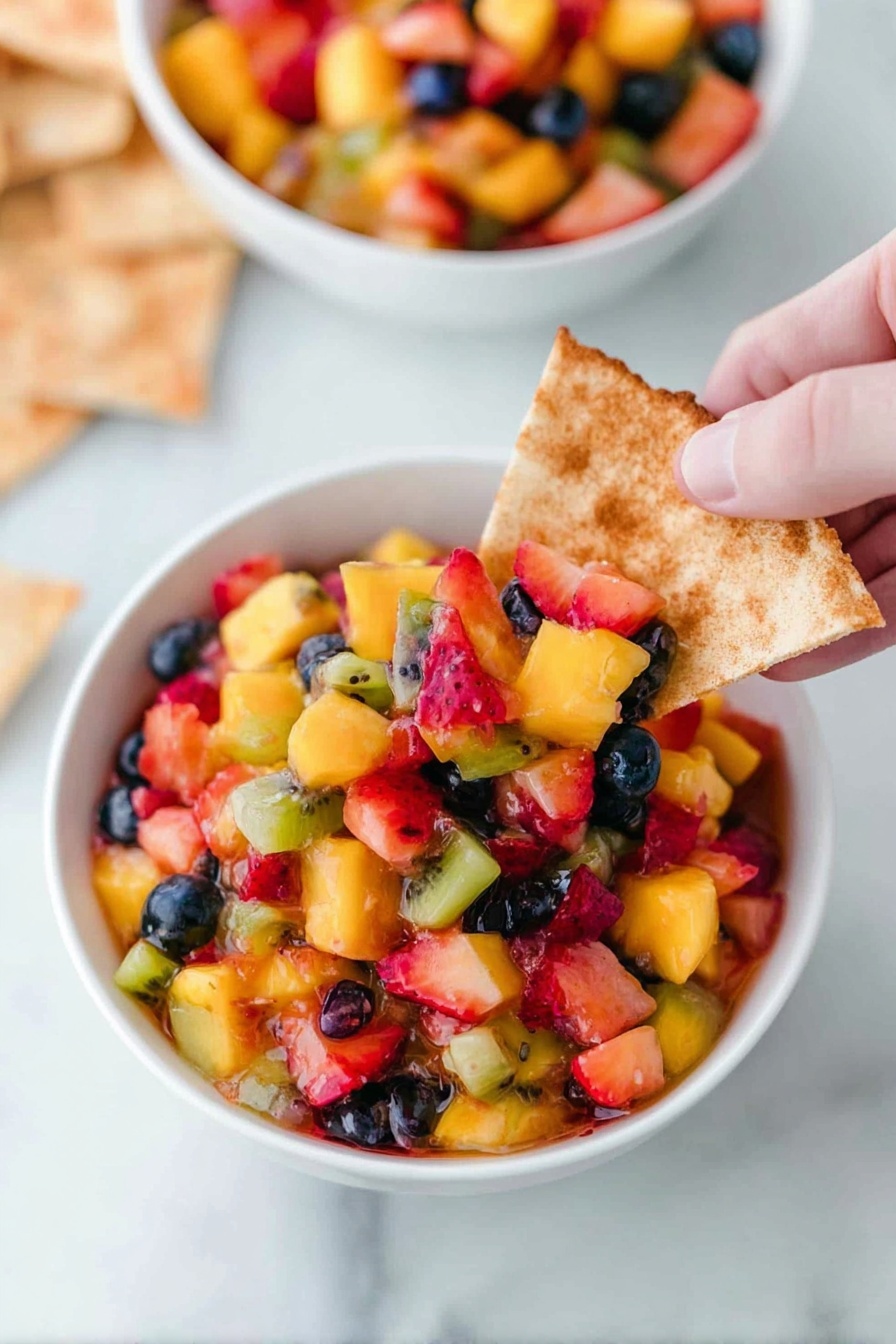 A white bowl filled to the top with a colorful fruit salsa made of small diced red strawberries, orange mango, light green kiwi, and whole dark blue blueberries. The fruit pieces are mixed evenly, creating a vibrant, juicy texture that shines under the light. The bowl sits on a white plate surrounded by triangular toasted pita chips that are light brown with a cinnamon-like dusting on top. The background is a white marbled surface, adding a clean and bright feel to the image. photo taken with an iphone --ar 2:3 --v 7