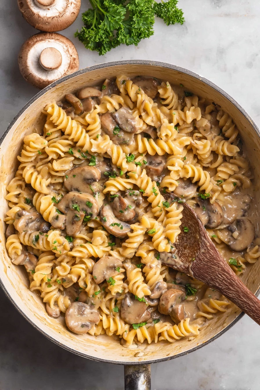 Vegan Mushroom Stroganoff - The image shows a close-up of a white deep pan filled with creamy mushroom pasta. The pasta is spiral-shaped and has a yellowish color, mixed well with sliced light brown mushrooms. The sauce is thick and creamy, coating the pasta and mushrooms evenly. Small pieces of green parsley are sprinkled on top, adding specks of color. A wooden spoon is resting in the pan on the right side, partially submerged in the pasta. The pan is placed on a white marbled surface. photo taken with an iphone --ar 2:3 --v 7