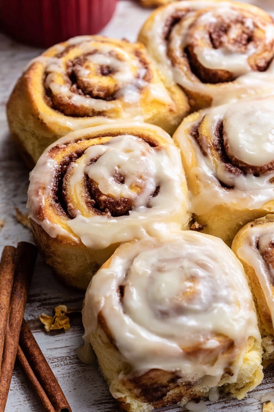 A close-up view of a group of warm cinnamon rolls on a white marbled surface, arranged in rows on parchment paper; each roll has a thick swirl pattern with golden brown dough and darker cinnamon filling visible inside, all topped with a generous layer of creamy white icing that is smooth and slightly shiny, some icing dripping down the sides; a silver spoon with icing rests nearby with more cinnamon sticks scattered around and a white pitcher with a red rim partially visible above the rolls; the top right shows a woman's hand holding one cinnamon roll slightly lifted from the group photo taken with an iphone --ar 2:3 --v 7