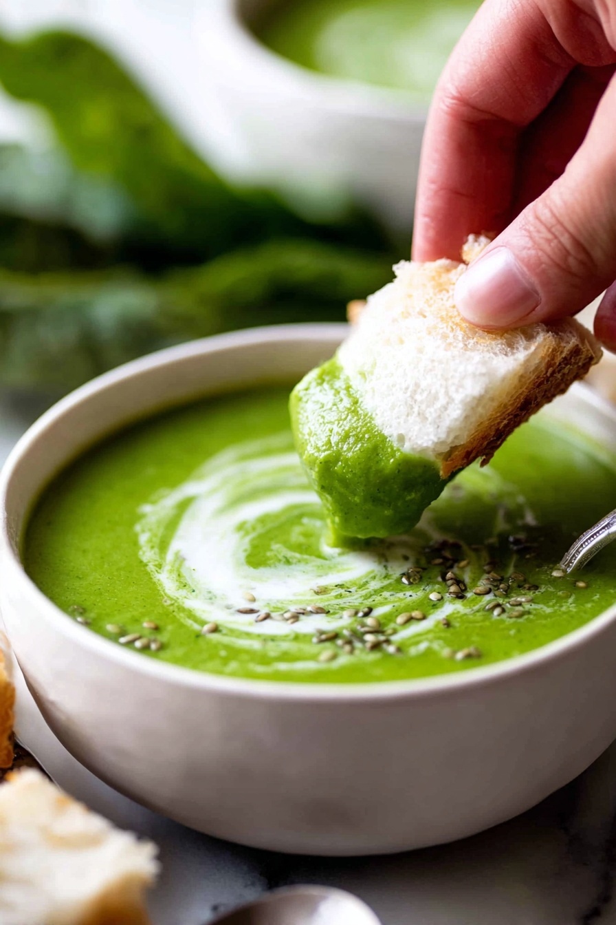A close-up view of a bowl filled with smooth, vibrant green soup with a slight creamy swirl on top, garnished with small seeds. A woman's hand is dipping a piece of soft, white bread into the soup, showing the bread partially covered in the green soup and cream. The bowl is white, placed on a white marbled surface with blurred green leaves in the background. Photo taken with an iphone --ar 2:3 --v 7