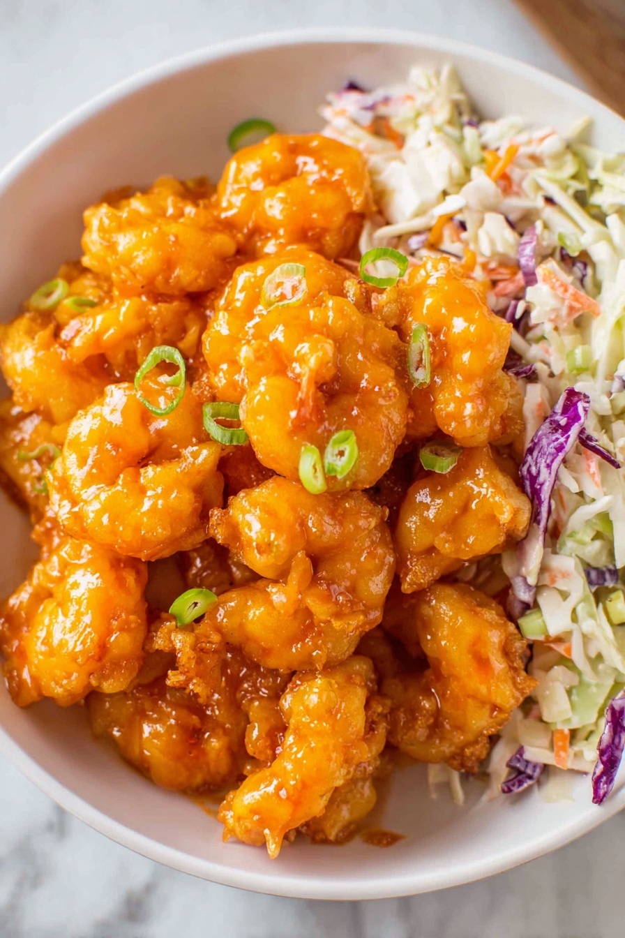 A white bowl filled with bright orange fried shrimp coated in a shiny sauce, piled high in the center. On the top right side of the bowl, there is a layer of chopped coleslaw with white, purple, and orange pieces, adding a fresh, crunchy texture. Small green scallion slices are sprinkled on top of the shrimp, giving a pop of color and freshness. The bowl sits on a white marbled surface, highlighting the vibrant colors of the food. photo taken with an iphone --ar 2:3 --v 7