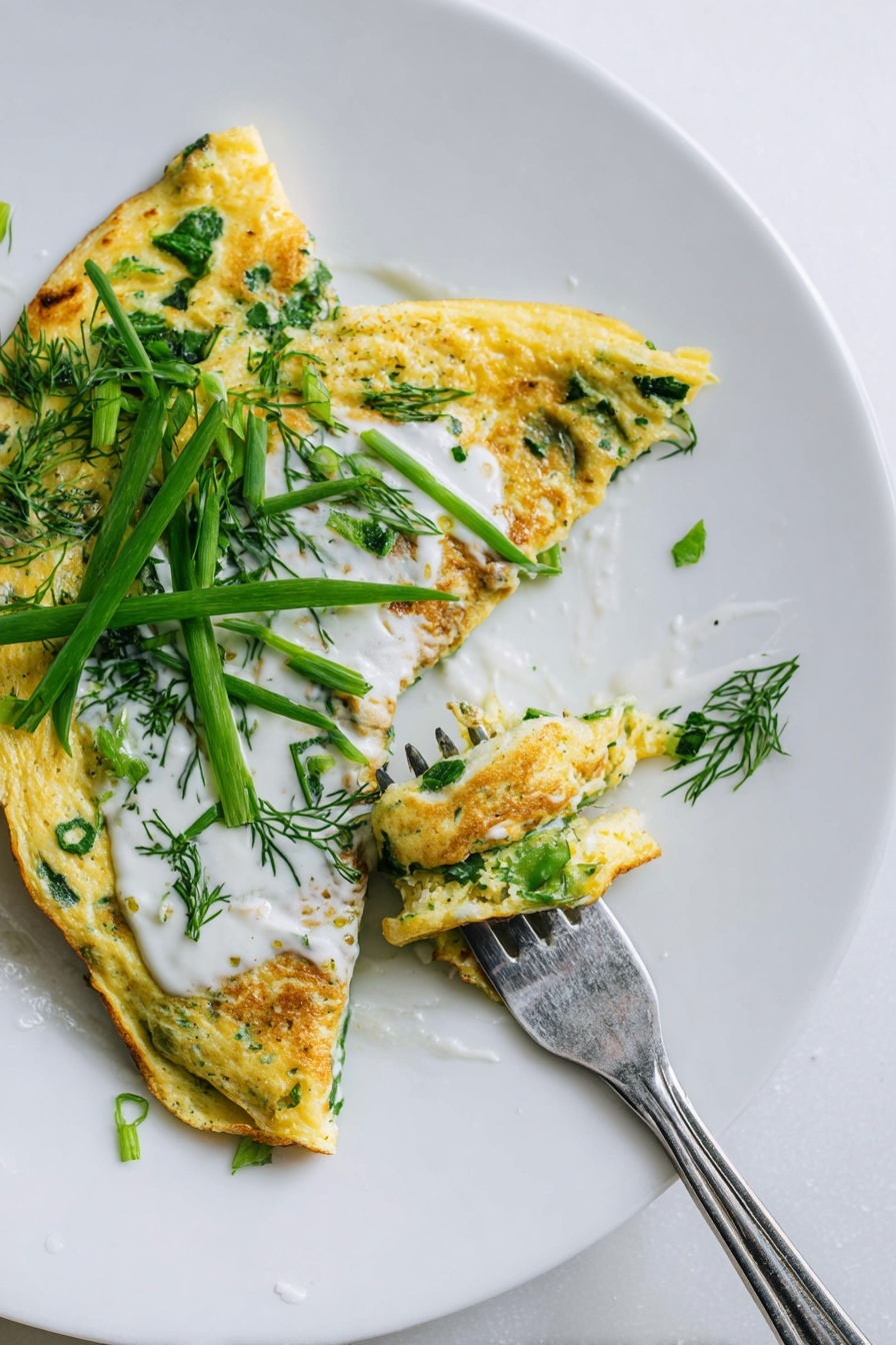 A white plate on a white marbled surface holds a folded yellow omelette speckled with green herbs and scallions. The omelette is topped with a smooth white sauce spread along part of it, and fresh green herbs and scallion strips rest on top. A piece of the omelette is cut and held by a fork resting on the plate’s edge. Photo taken with an iphone --ar 2:3 --v 7