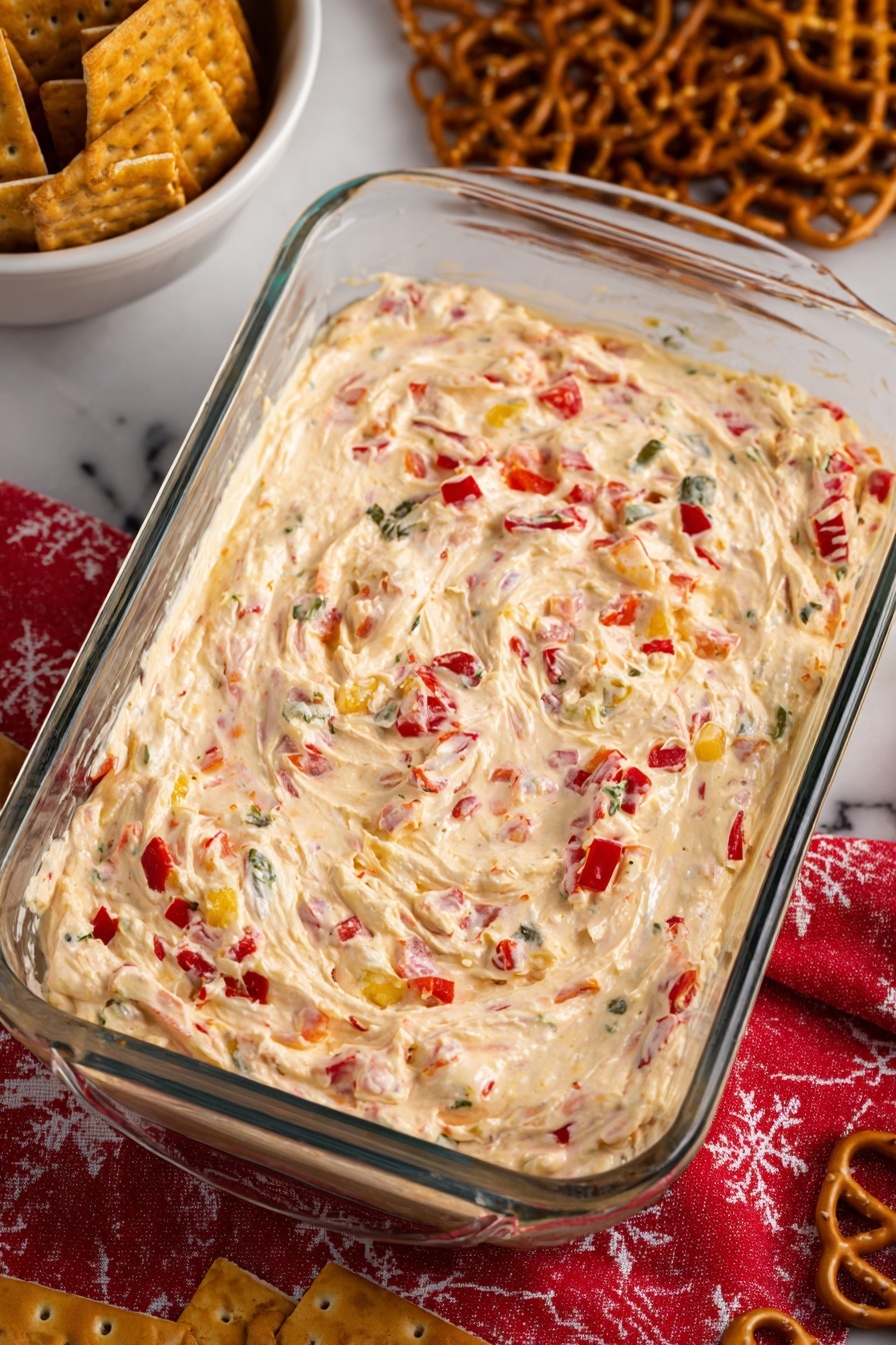 The dish is a thick, creamy mixture with a swirled surface, set inside a clear glass rectangular container. The base is light cream color, dotted generously with small pieces of red and green, which look like chopped peppers or vegetables. The texture appears smooth but dense, with visible soft lumps throughout. Around the container, a white marbled surface is visible, and a red cloth with a white pattern is placed underneath. To the top left, there is a white bowl filled with brown square crackers, and some lattice-shaped brown pretzels are scattered on the right side. Photo taken with an iphone --ar 2:3 --v 7