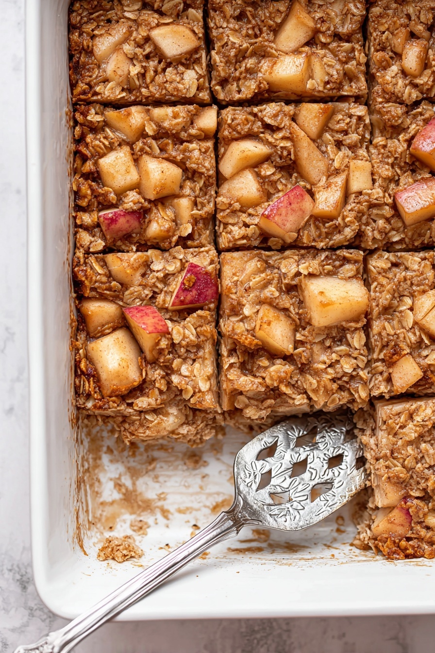 A square piece of baked oatmeal sits on a white scalloped plate with syrup pooled around its base. The oatmeal bar shows a dense texture with visible oats and small apple chunks inside, all in golden-brown shades. On top, three thin slices of red-skinned apple are laid in a small stack at an angle. Next to the plate is a silver fork and in the background, a white pitcher and a glass bottle with milk sit on a white marbled surface. photo taken with an iphone --ar 2:3 --v 7