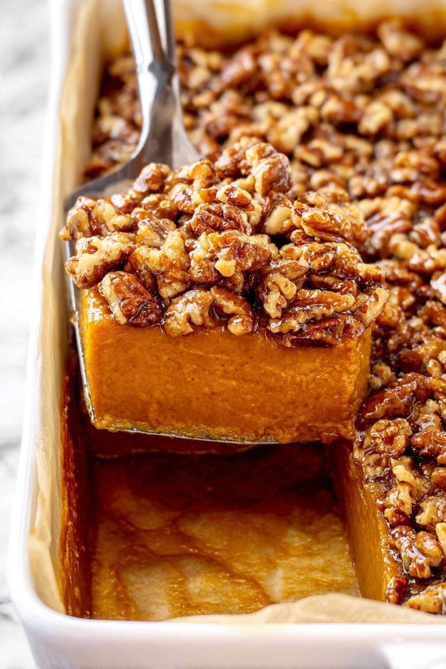 This image shows a dessert with two clear layers in a white rectangular dish lined with parchment paper. The bottom layer is smooth, thick, and deep orange-brown, resembling a pumpkin custard. The top layer is made up of chopped pecans, golden brown and glossy, adding a textured crunch. A metal spatula is lifting a piece, showing the contrast between the creamy orange-brown base and the nutty top. The background is a white marbled texture. photo taken with an iphone --ar 2:3 --v 7