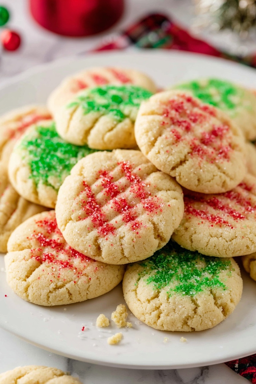 A white plate holds about fifteen soft round cookies stacked in a loose pile, each cookie a pale golden color with a slightly cracked surface. Each cookie is decorated on top with coarse sugar sprinkles in two colors: bright red and bright green, scattered mostly in the center and fading outwards. Each cookie has a shallow grid pattern pressed into its top, made by pressing with a fork, giving it a textured look. Crumbs and a few sprinkles scatter lightly around the plate edge. The background is a white marbled surface and blurred holiday items add soft colors behind the plate. Photo taken with an iphone --ar 2:3 --v 7