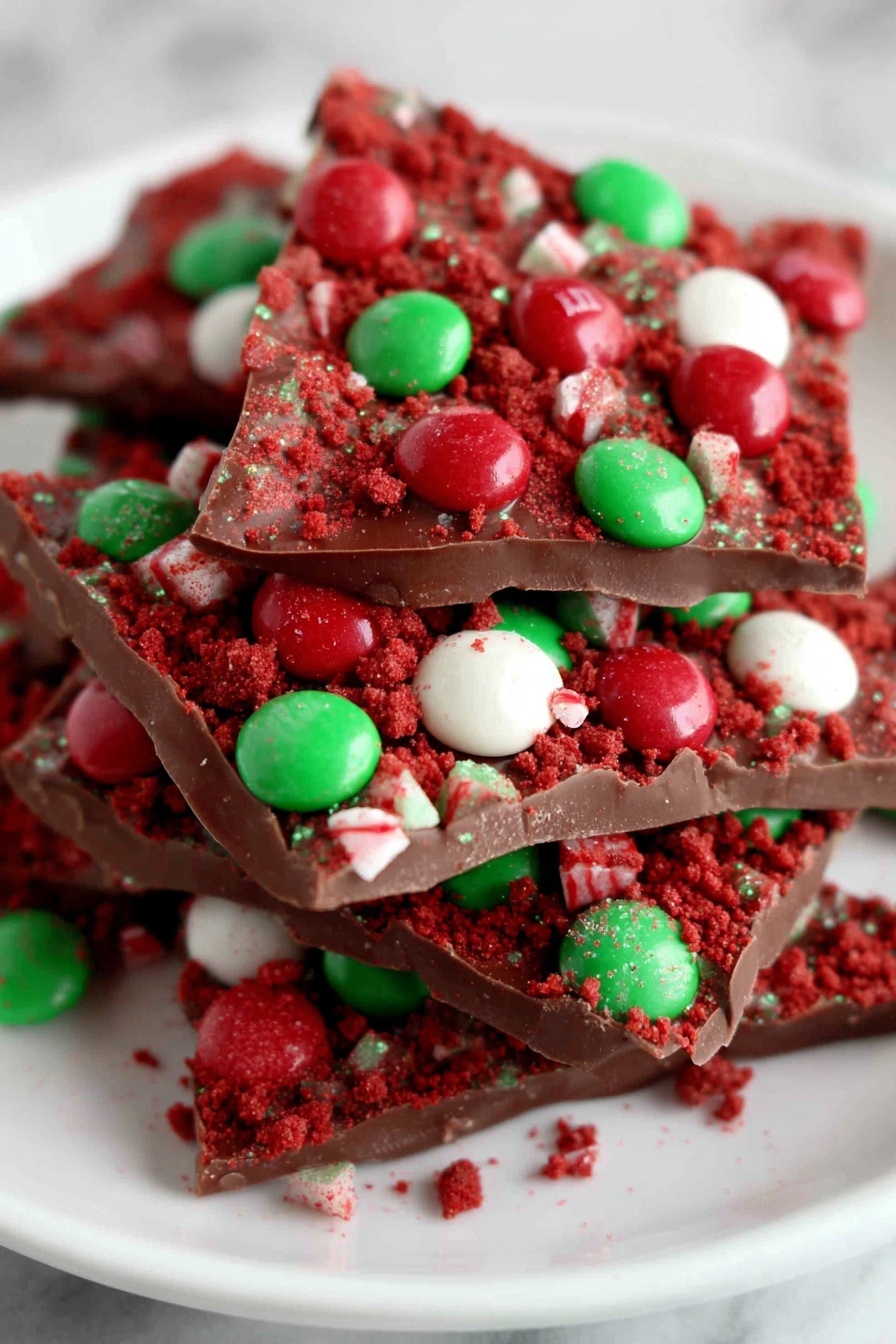 The image shows several pieces of chocolate bark stacked on a white plate over a white marbled surface. Each piece has a smooth, dark brown chocolate base topped with crushed red cake crumbs and whole red, green, and white candy-coated chocolates spread unevenly across the surface. The candy pieces stand out with their bright colors contrasting with the chocolate and red crumbs, creating a festive look. photo taken with an iphone --ar 2:3 --v 7