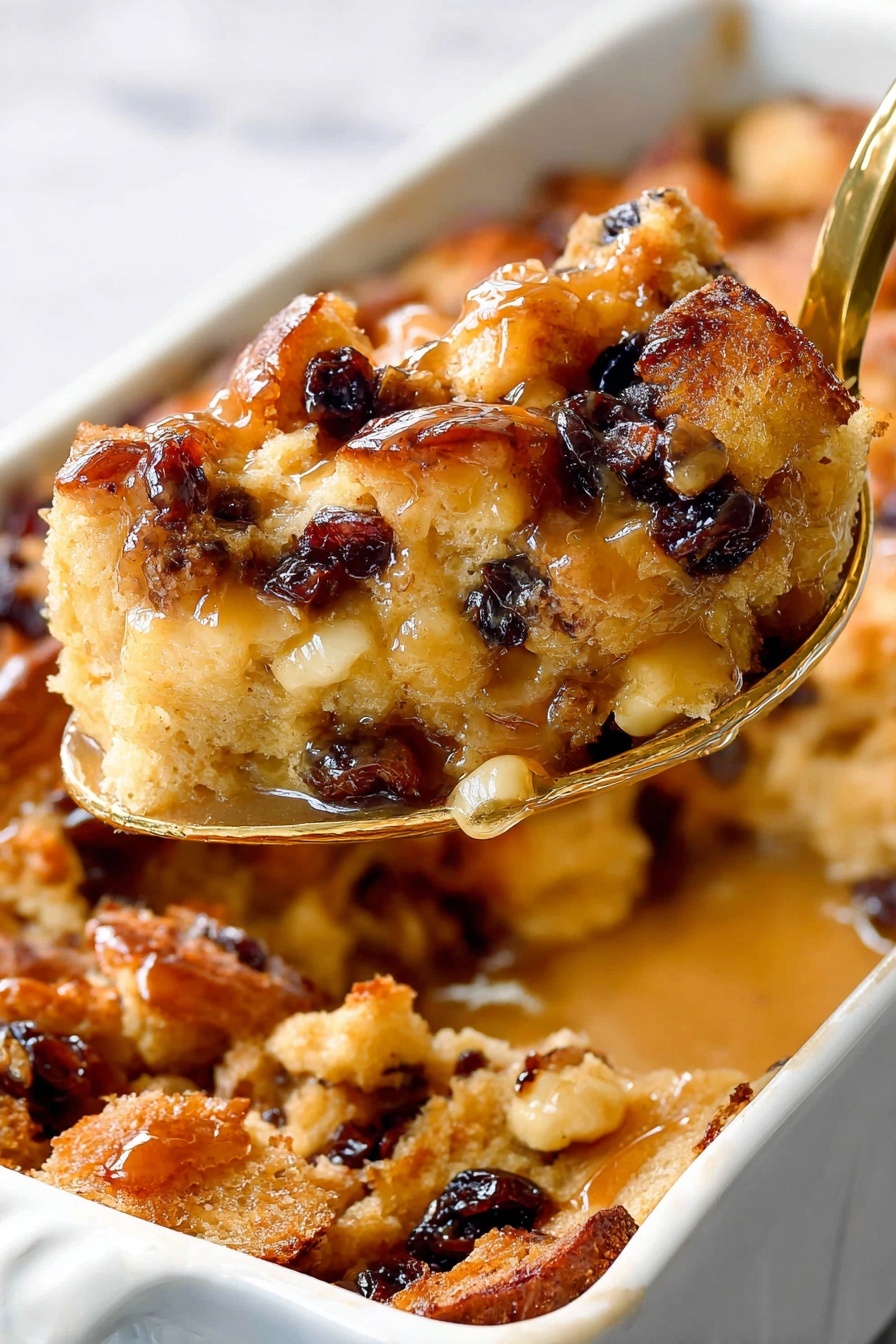 The image shows a rectangular white baking dish filled with a bread pudding-like dessert. The dish contains several layers of uneven golden-brown bread chunks soaked in a shiny, light brown sauce. Mixed inside are small dark dried fruits and small pieces of pale yellow fruit, creating a mottled texture on the surface. The edges of the pieces are slightly crispy, while the liquid sauce adds a glossy look, making the dessert appear moist and rich. The dish is set on a white marbled surface. photo taken with an iphone --ar 2:3 --v 7