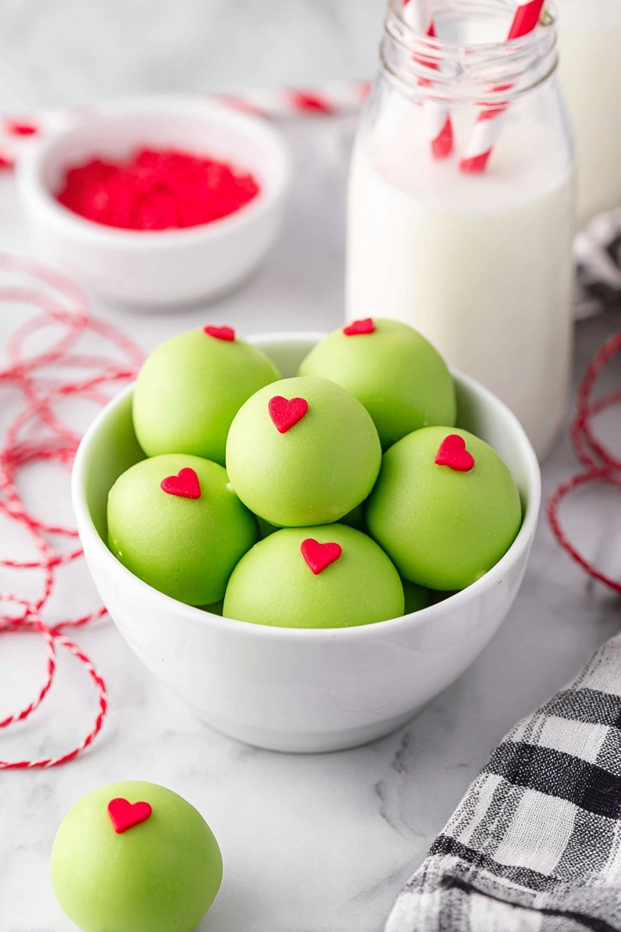 A white bowl filled with six round green cake balls, each smooth and covered with a light green coating. Each cake ball has a small red heart decoration placed on top at the center. One cake ball sits outside the bowl on a white marbled surface. In the background, there is a glass bottle of milk with two red and white striped straws, and a white bowl with red heart sprinkles. A red and white twisted string and a black and white checkered cloth are also visible on the white marbled surface. photo taken with an iphone --ar 2:3 --v 7