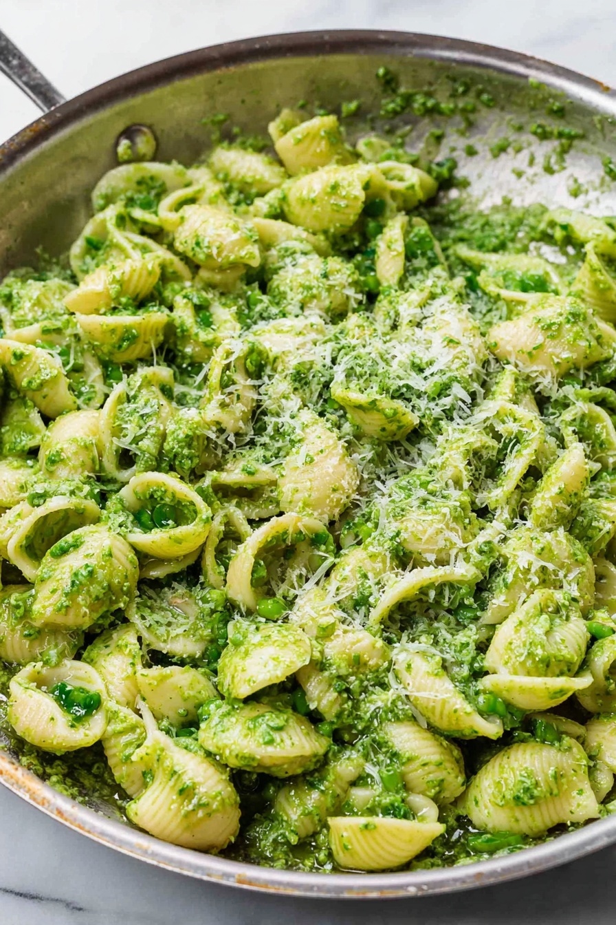 A silver pan filled with a large amount of small white shell pasta covered in a bright green sauce, likely pesto, with a slightly chunky texture. The pasta shells are well coated and mixed, and there is a sprinkling of fine white grated cheese on top, adding a light contrast to the green. The background shows a white marbled surface, and the pan’s edges are slightly worn. The image is clear and close up, showing the texture of the pasta and sauce in detail photo taken with an iphone --ar 2:3 --v 7