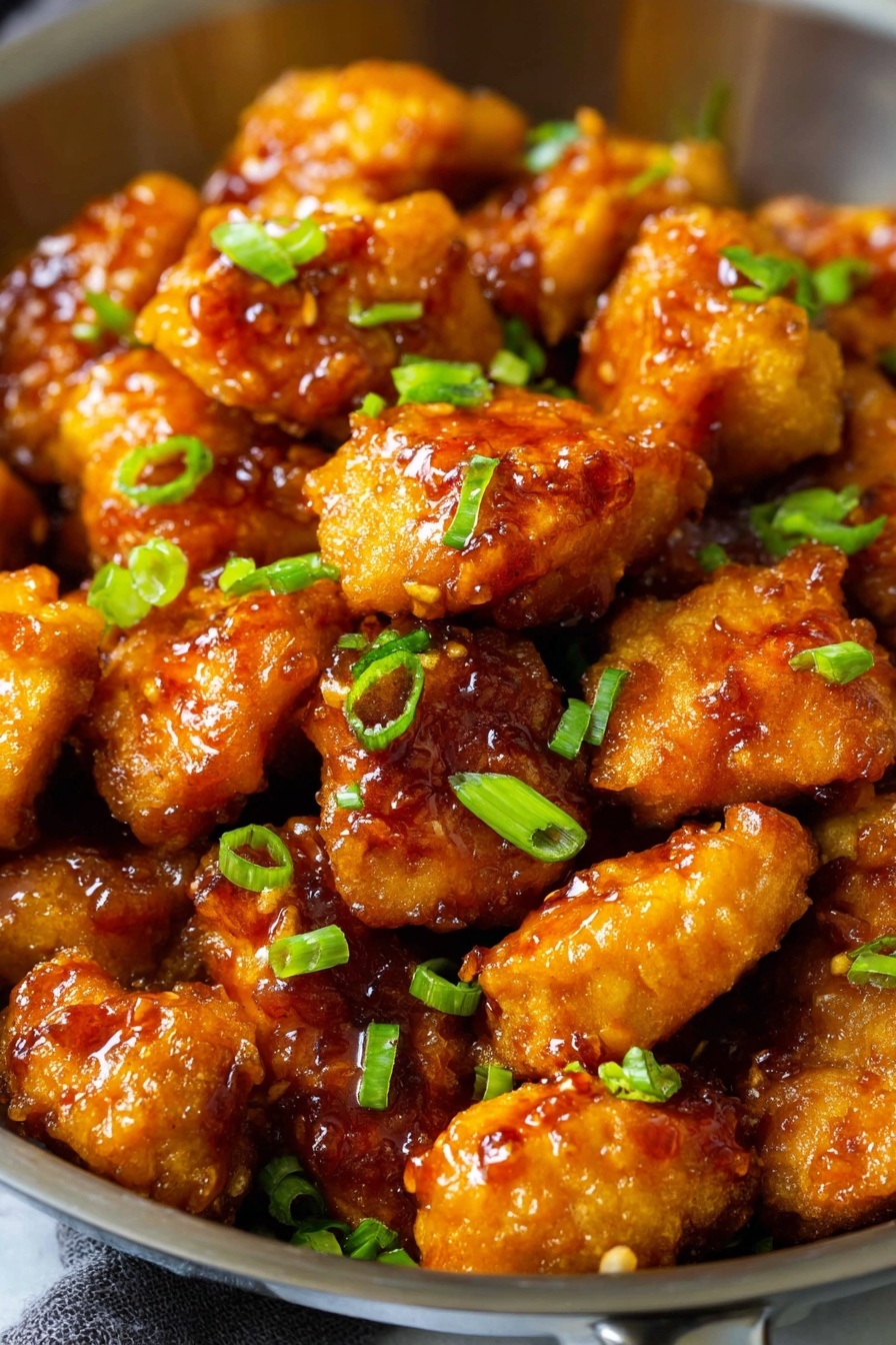The image shows a close-up of a pan filled with multiple golden-brown fried nuggets covered in a shiny orange glaze. Each nugget has a crispy texture and is topped with small pieces of fresh green onion scattered evenly across them. The nuggets have a slight sheen from the sticky glaze, which looks thick and rich, adding a glossy finish. The pan is silver-colored, and the background is a white marbled texture. photo taken with an iphone --ar 2:3 --v 7