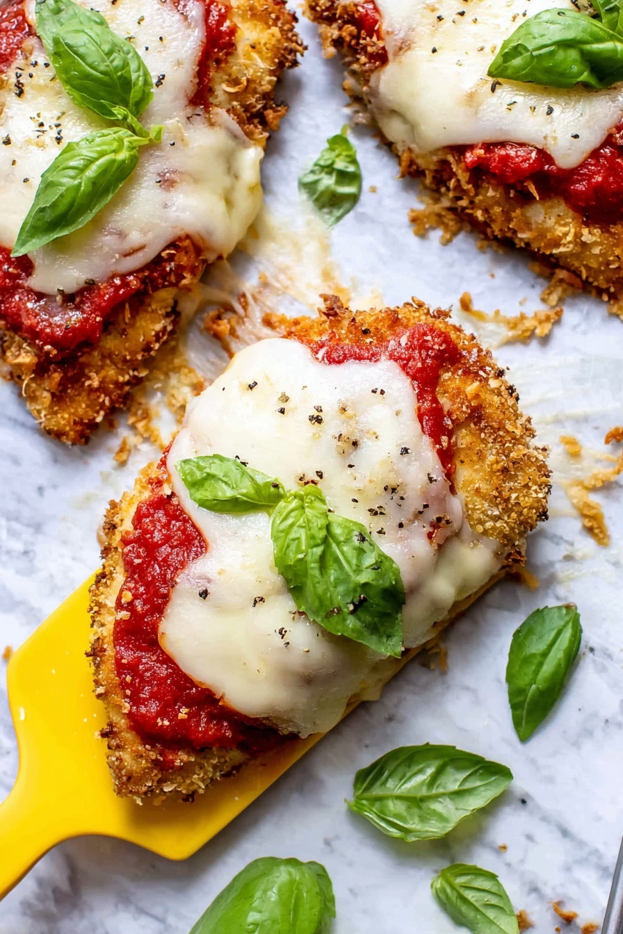 The image shows three pieces of breaded and cooked chicken topped with a layer of bright red tomato sauce, followed by a thick layer of melted white cheese. Each piece is garnished with a fresh green basil leaf on top. The chicken pieces have a golden-brown crispy texture with bits of crumbs scattered around them. A yellow spatula is lifting the front piece on a white marbled surface, with more basil leaves spread around for decoration. The melted cheese has a stretchy, melting texture with some pepper sprinkled on top. Photo taken with an iphone --ar 2:3 --v 7