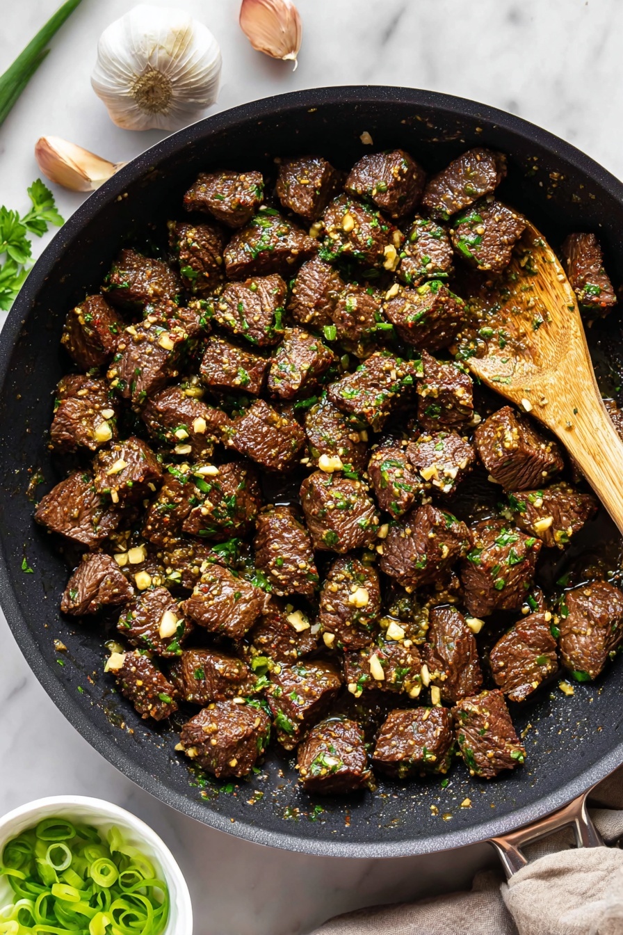 The image shows a black frying pan filled with many small, dark brown cooked beef cubes that have a slightly shiny, moist surface. Each beef cube is covered with tiny pieces of light-colored cooked garlic and small green herb leaves, giving a fresh and textured look. In the pan, near the top right, there is a wooden spoon resting among the beef cubes. The pan is placed on a white marbled surface with three garlic cloves near the top left and some green parsley leaves at the edge. A small white bowl filled with sliced green onions is partly visible at the bottom left. The lighting is natural and bright, showing the rich colors and textures clearly. Photo taken with an iphone --ar 2:3 --v 7