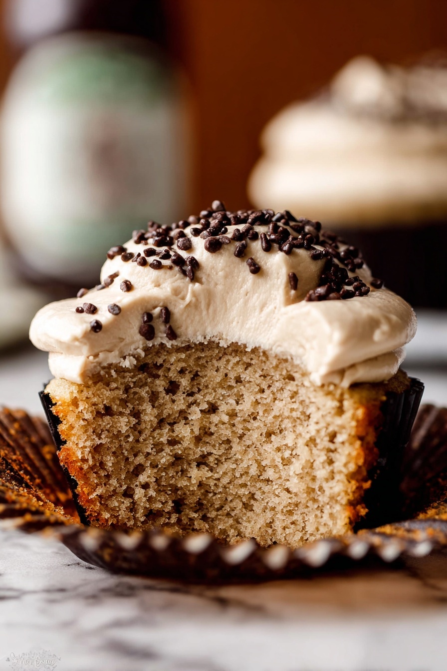 A close-up view of a thick creamy light beige frosting swirled inside a metal mixing bowl. The frosting has a smooth, soft texture with visible folds and peaks, and a red spatula is partially dipped in the frosting on the right side. The bowl sits on a white marbled surface with a striped brown cloth partially visible underneath. photo taken with an iphone --ar 2:3 --v 7