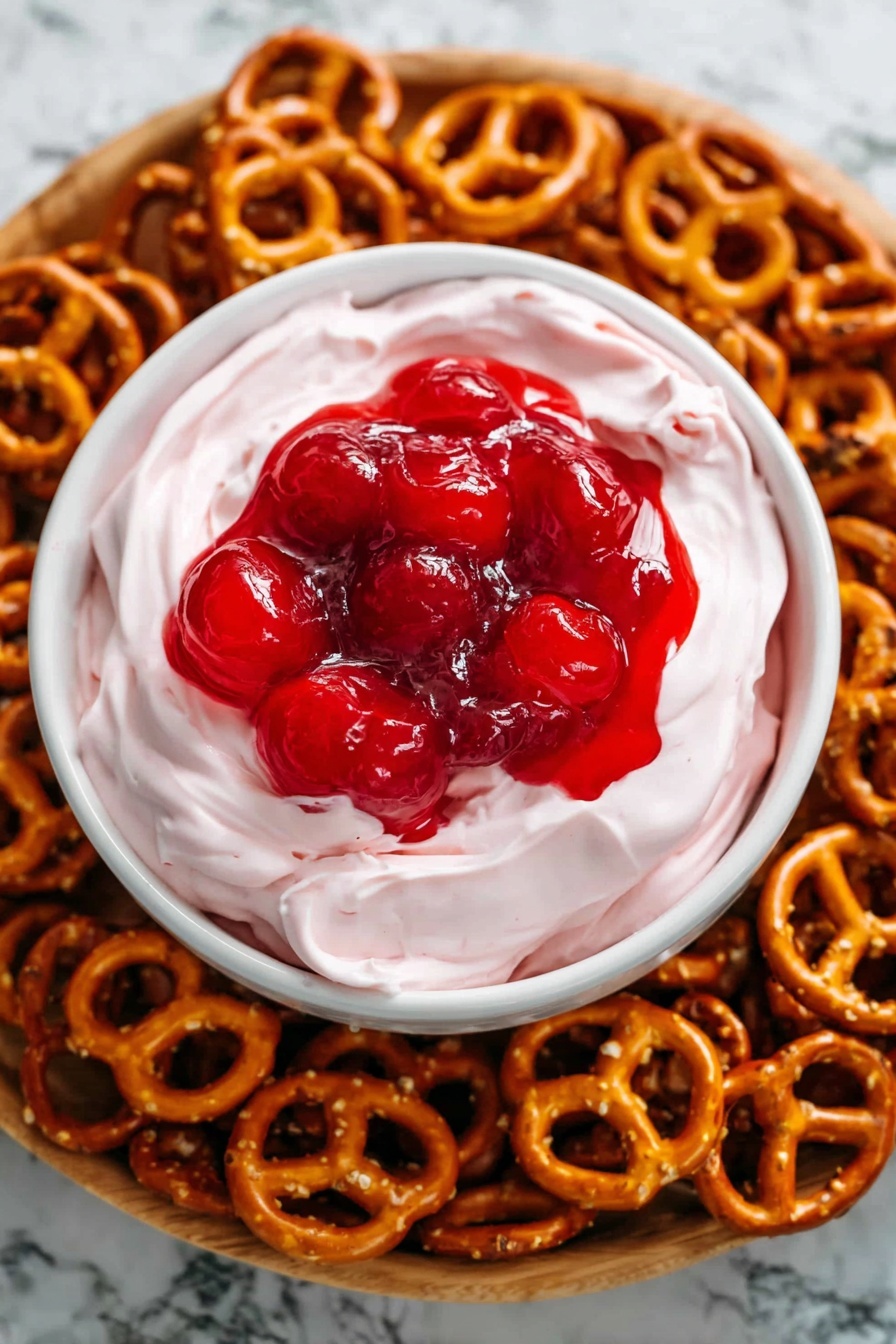 A white bowl with two layers sits in the center of a wooden tray filled with small golden brown pretzels. The first layer in the bowl is a smooth, light pink creamy dip that looks thick and soft, filling most of the bowl. On top of this dip is a glossy bright red cherry topping with whole cherries visible, creating a shiny, slightly wet texture. The wooden tray underneath is light brown and has a wood texture background. photo taken with an iphone --ar 2:3 --v 7