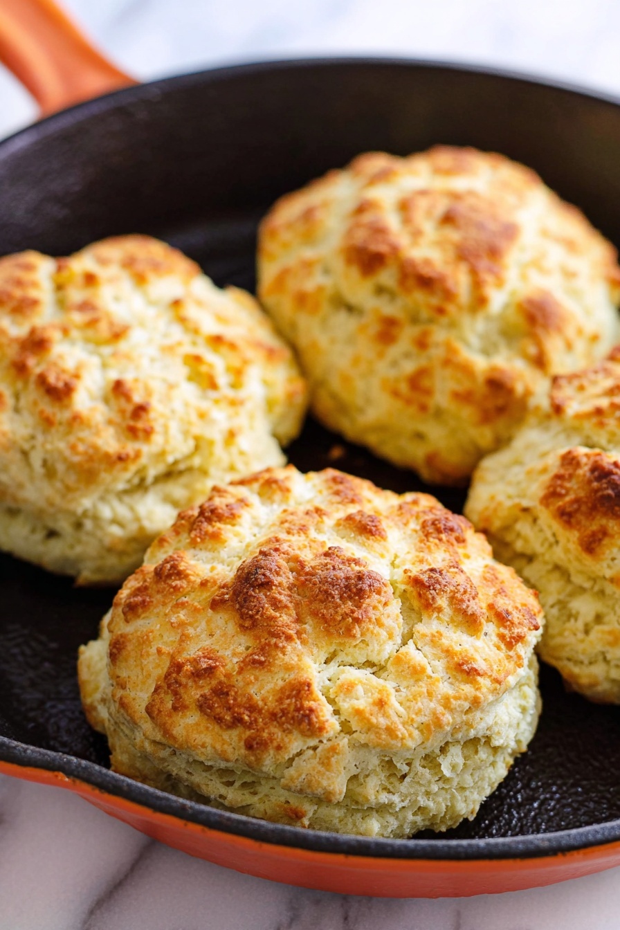 The image shows two cast iron pans with five round dough pieces in each, arranged with one in the center and four around it. The left pan holds raw dough balls that are pale and rough in texture with a white color, while the right pan displays the same dough pieces baked to a golden brown with a slightly crispy and uneven surface. Both pans are black with bright orange handles, placed on a white marbled surface that has subtle gray vein patterns. The contrast between the raw and baked biscuits highlights their texture and color change clearly. photo taken with an iphone --ar 2:3 --v 7